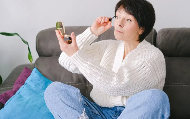 A senior caucasian woman sitting on a couch applying mascara in a cozy setting.