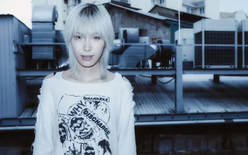 Stylish young adult with platinum hair stands against urban industrial backdrop.