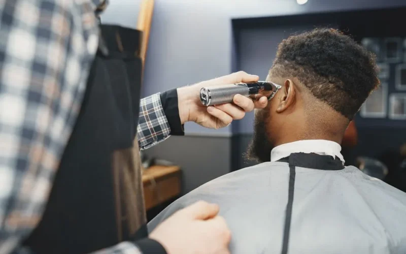 A barber uses clippers to style an African American man's hair in a salon setting.