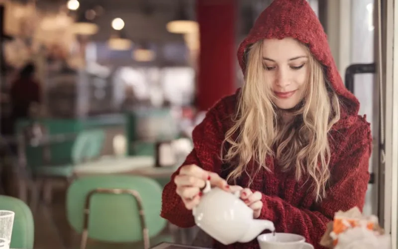 Blonde woman in red sweater pouring tea in a cozy cafe setting.