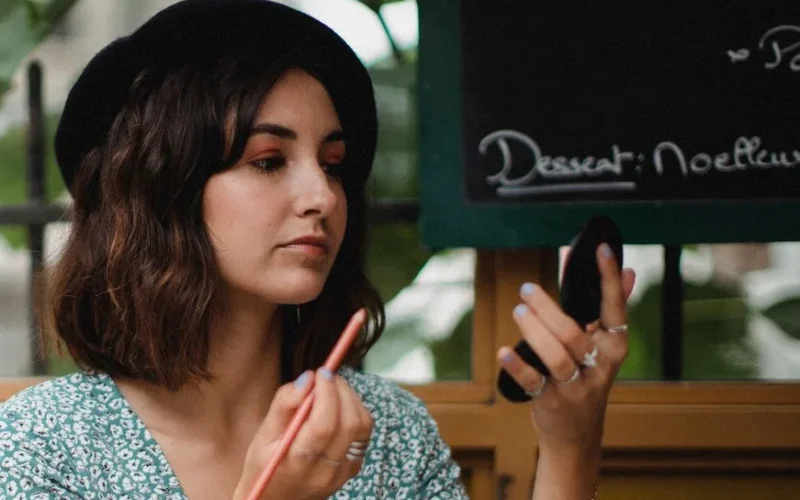 Young woman uses compact mirror for makeup touch-up in cozy café.