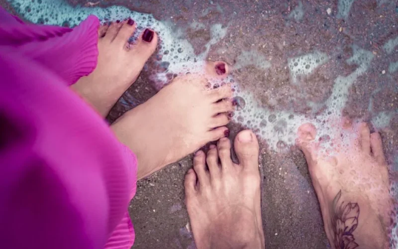 Three pairs of feet in ocean waves with sandy beach and purple attire.