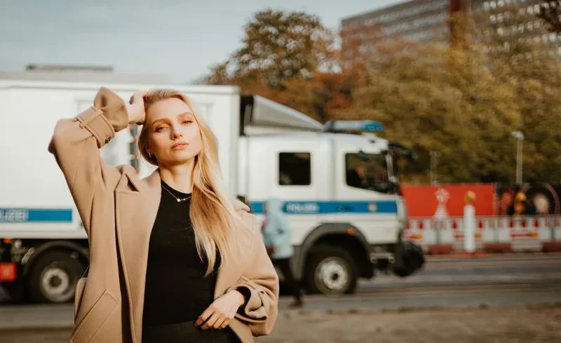 Stylish woman in coat posing on a city street with a truck in the background.