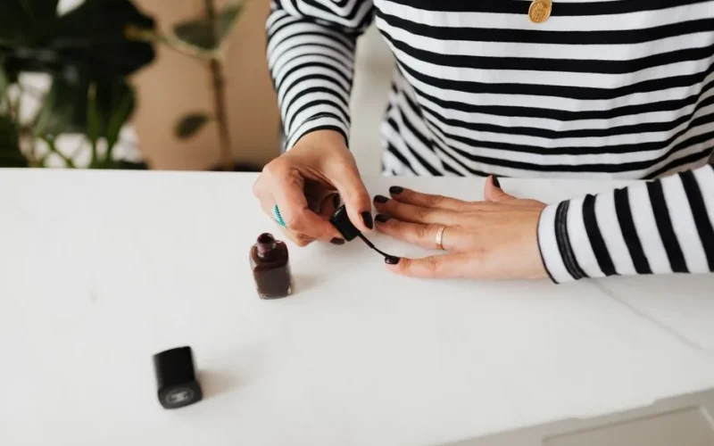 Unrecognizable female doing manicure with black nail polish on white table resting at home during weekend
