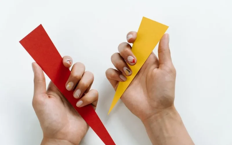 Two hands holding red and yellow geometric paper shapes on a minimalist background.