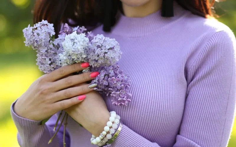 Woman's hands holding lilac flowers, wearing a lavender sweater with vibrant nails in sunlight.