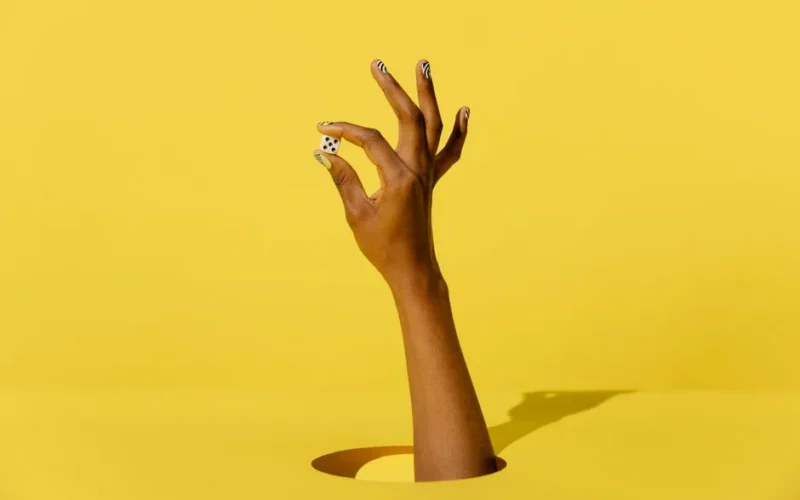 A hand with trendy nail art holds a dice on a vibrant yellow background, creating a modern and playful image.