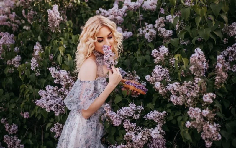 A blonde woman in a floral dress embraces the beauty of lavender blooms in a lush garden setting.