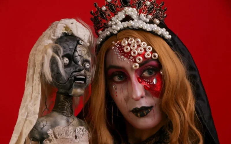 Artistic portrait of a woman with gothic Halloween makeup and a creepy doll against a red background.