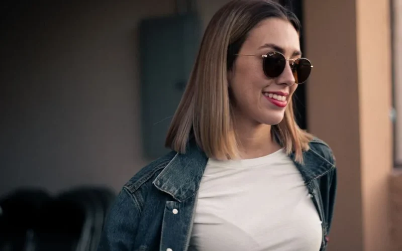 A woman with short blonde hair and sunglasses smiles outdoors wearing a denim jacket.