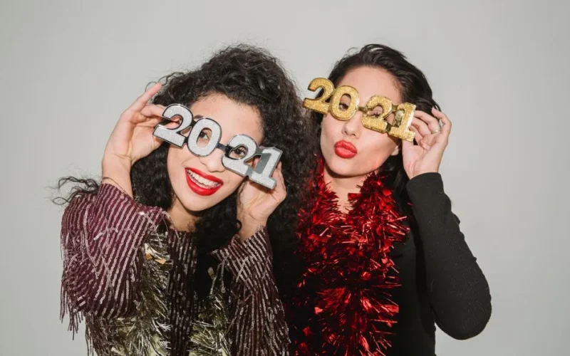 Cheerful women with bright red lips and sparkling red and golden tinsel looking at camera on gray background