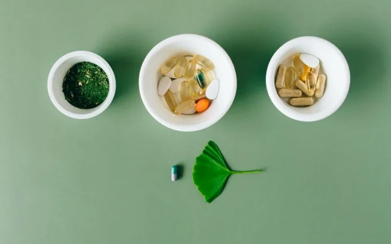 Flatlay of capsules and herbs in ceramic bowls on green surface.