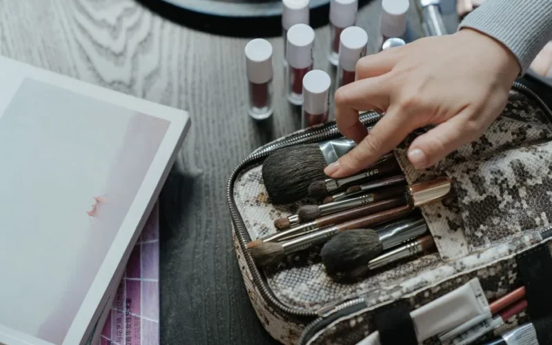 A hand arranging makeup brushes in a case surrounded by cosmetics in a flat lay setup.