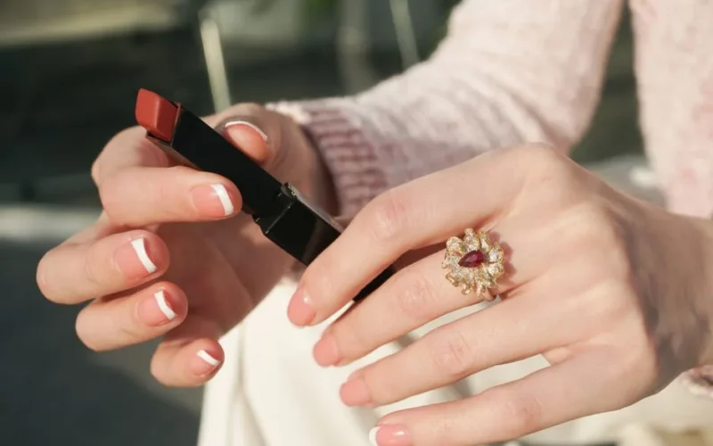 Elegant hands with French manicure holding lipstick, featuring a ruby ring in a sunny setting.