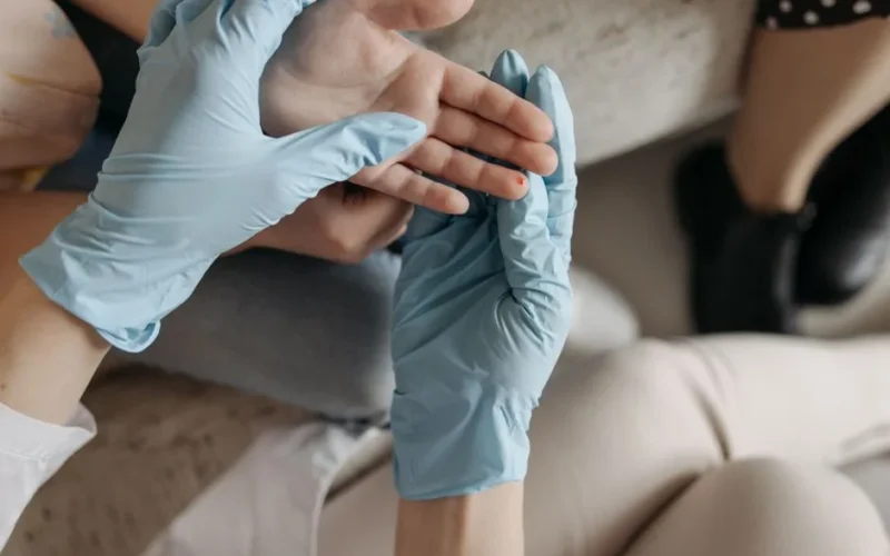 A medical professional wearing blue gloves examines a small wound on a person's finger indoors.