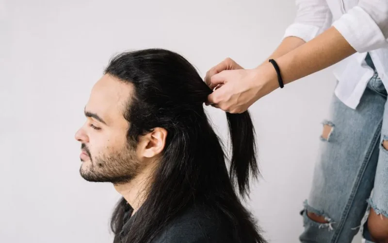 Profile view of a man with long hair being styled by a hairdresser indoors.